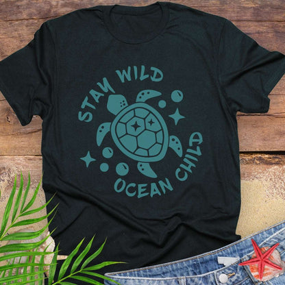 Black t-shirt with teal sea turtle graphic and 'Stay Wild Ocean Child' text on a wooden surface with leaves and jeans.