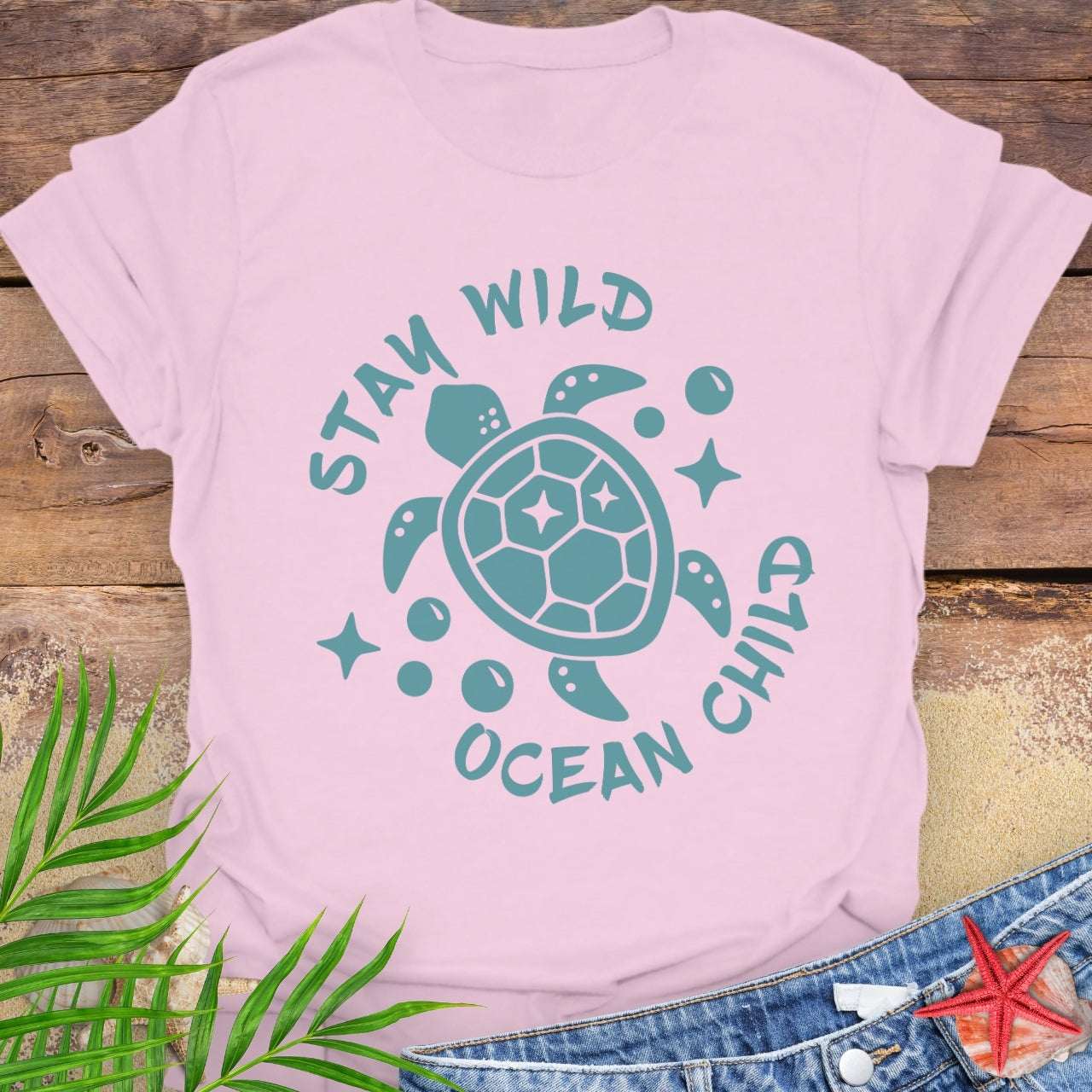 Pink t-shirt with teal turtle graphic and text 'Stay Wild Ocean Child' on a wooden surface with leaves and starfish.