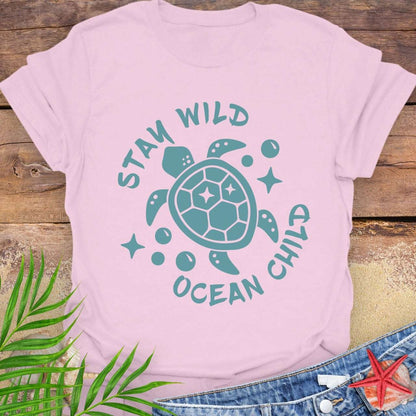 Pink t-shirt with teal turtle graphic and text 'Stay Wild Ocean Child' on a wooden surface with leaves and starfish.