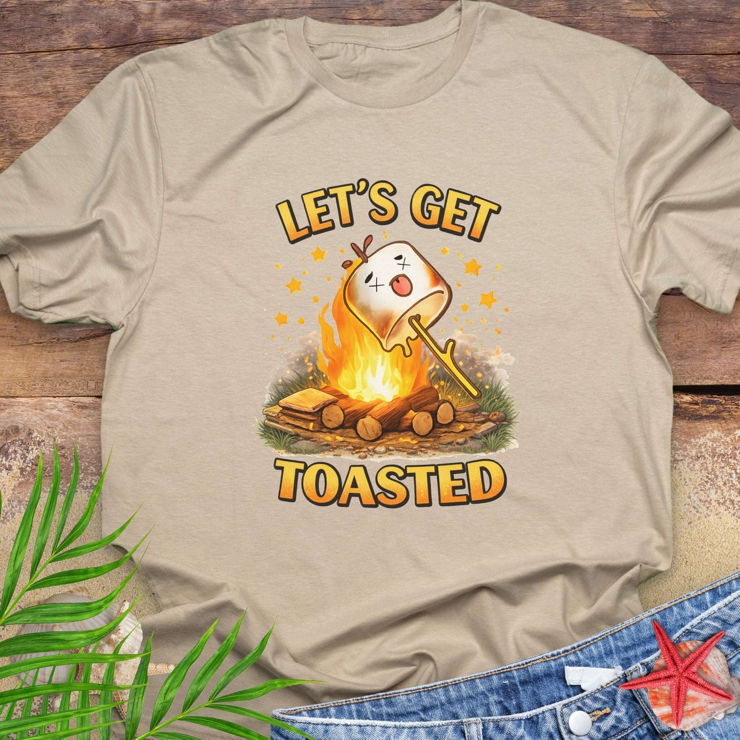 Beige t-shirt with a graphic of a marshmallow over a campfire and text 'Let's Get Toasted' on a wooden surface.