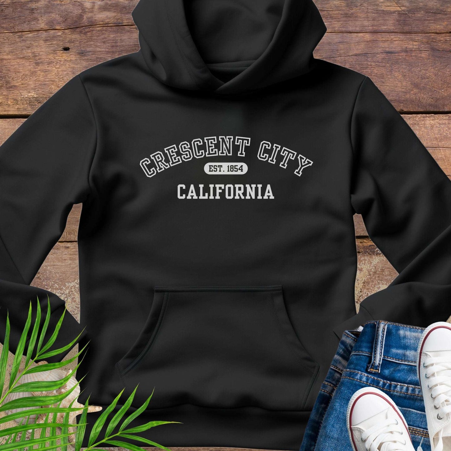 Black hoodie with 'Crescent City, California Est. 1854' text on a wooden surface with greenery and white sneakers.