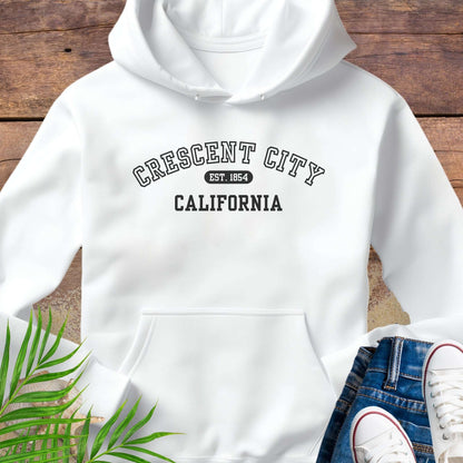 White hoodie with 'Crescent City, California EST 1854' text on a wooden surface with green leaves and white sneakers.