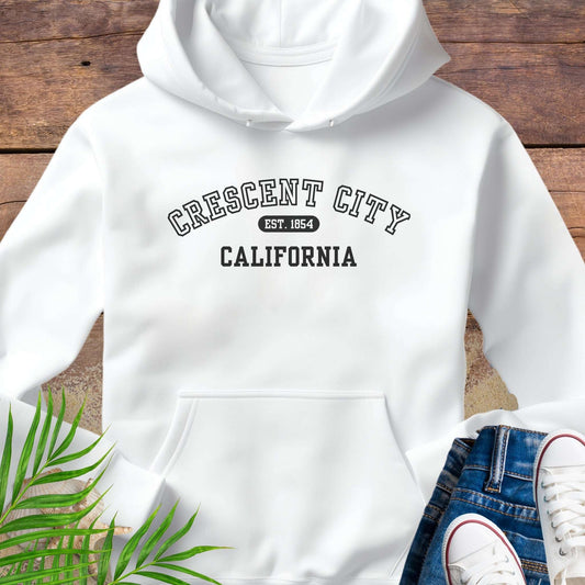 White hoodie with 'Crescent City, California EST 1854' text on a wooden surface with green leaves and white sneakers.