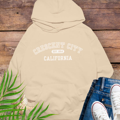 Beige hoodie with 'Crescent City, California Est. 1854' text on a wooden surface with greenery and white sneakers.