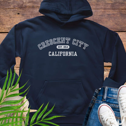 Navy blue hoodie with 'Crescent City, California Est. 1854' text on a wooden surface with greenery and white sneakers.