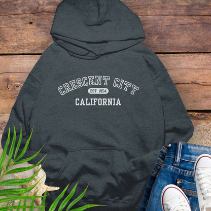 Dark gray hoodie with 'Crescent City, California Est. 1854' text on a wooden surface with greenery and white sneakers.