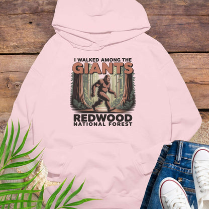 Pink hoodie with 'I Walked Among the Giants' design on a wooden surface with jeans and sneakers.