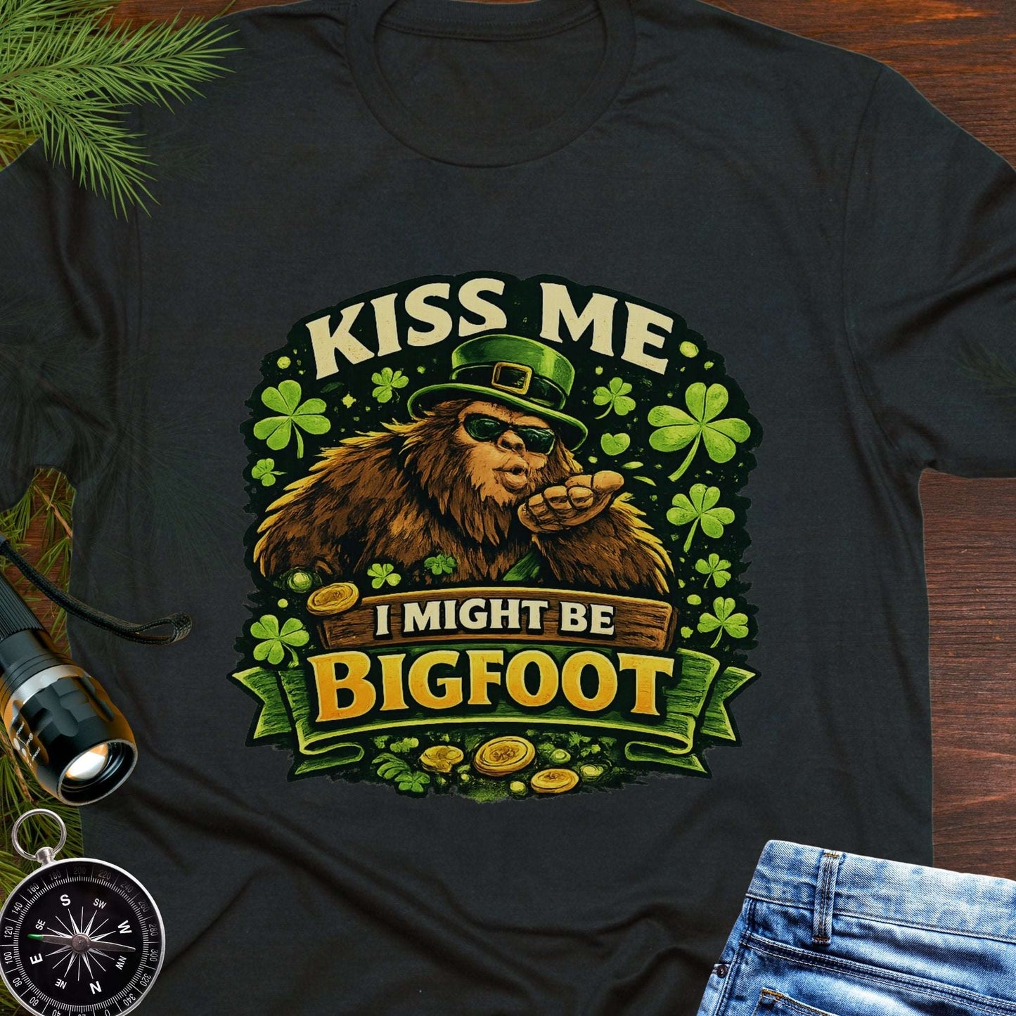 Black t-shirt with Bigfoot illustration wearing a leprechaun hat, surrounded by shamrocks and gold coins, with the text “Kiss Me, I Might Be Bigfoot”.