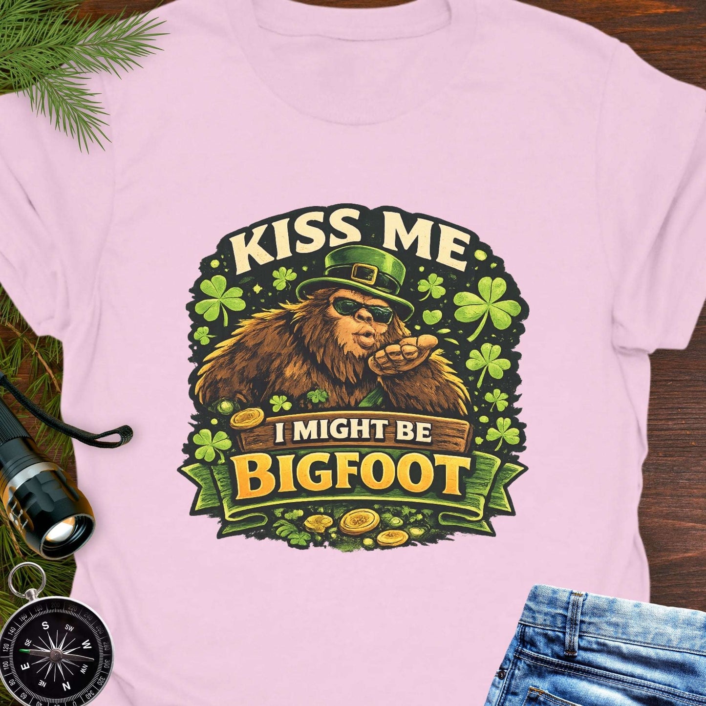 Pink t-shirt with Bigfoot illustration wearing a leprechaun hat, surrounded by shamrocks and gold coins, with the text “Kiss Me, I Might Be Bigfoot”.