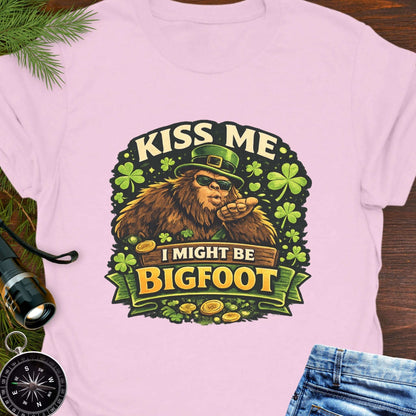 Pink t-shirt with Bigfoot illustration wearing a leprechaun hat, surrounded by shamrocks and gold coins, with the text “Kiss Me, I Might Be Bigfoot”.