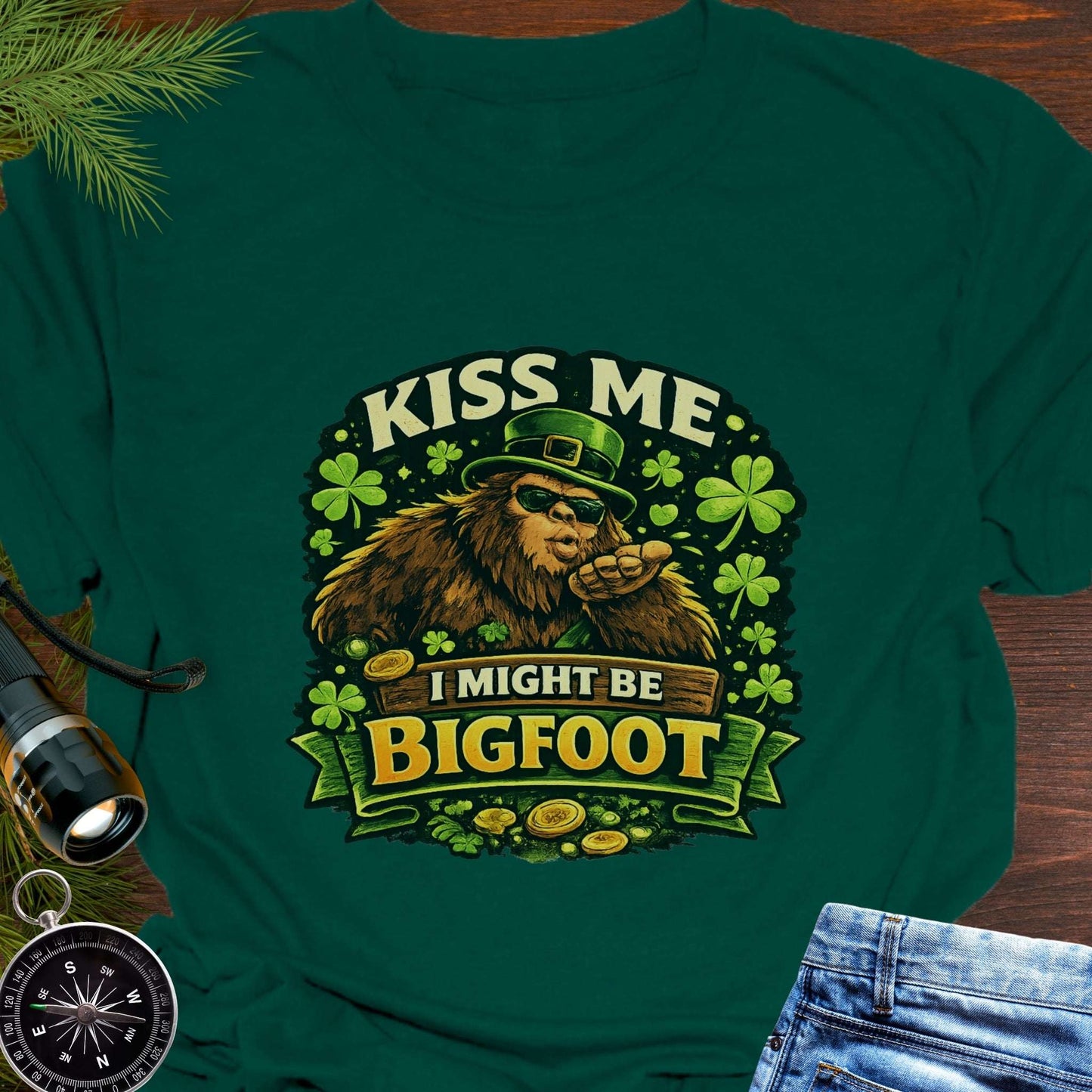Green t-shirt with Bigfoot illustration wearing a leprechaun hat, surrounded by shamrocks and gold coins, with the text “Kiss Me, I Might Be Bigfoot”.