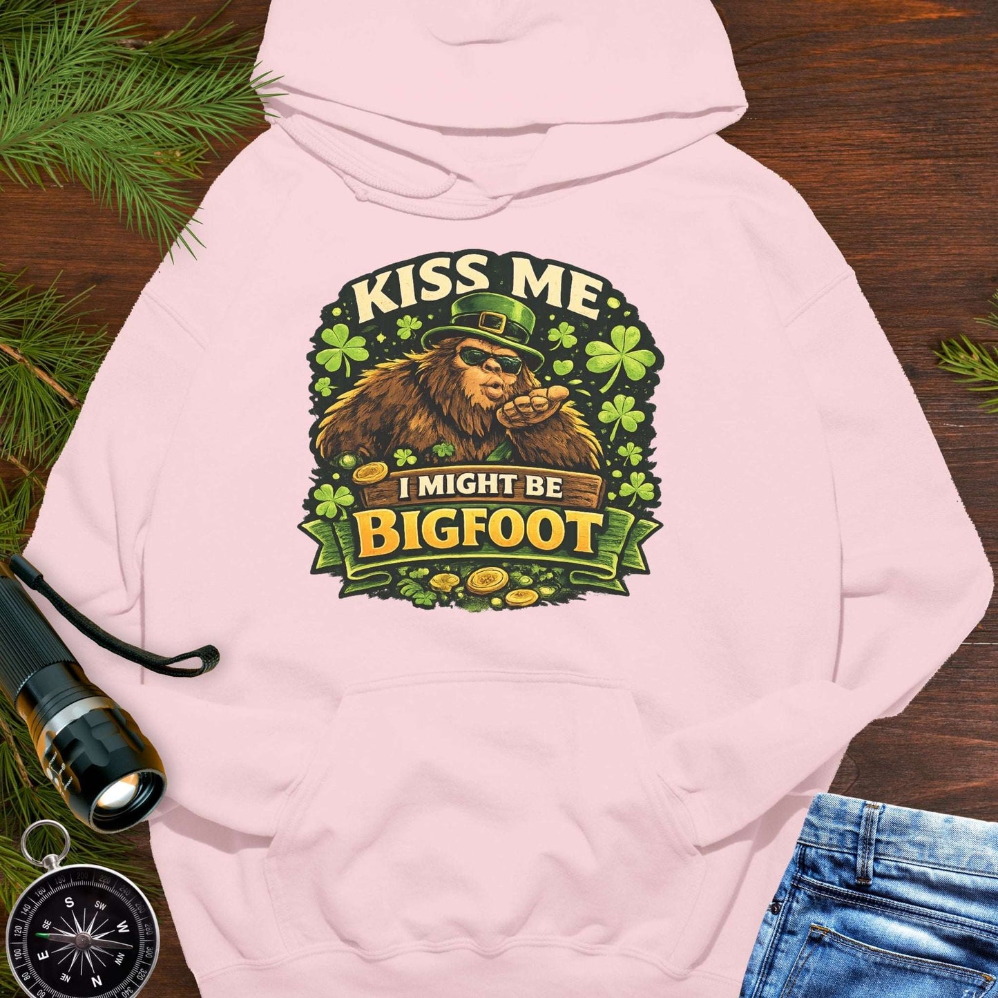 Pink hoodie with Bigfoot blowing a kiss, surrounded by shamrocks and gold coins, wearing a leprechaun hat, with the text “Kiss Me, I Might Be Bigfoot”.