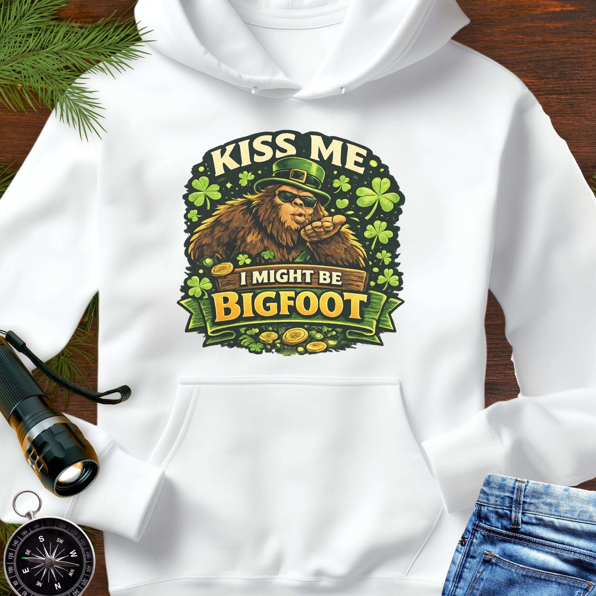 White hoodie with Bigfoot blowing a kiss, surrounded by shamrocks and gold coins, wearing a leprechaun hat, with the text “Kiss Me, I Might Be Bigfoot”.
