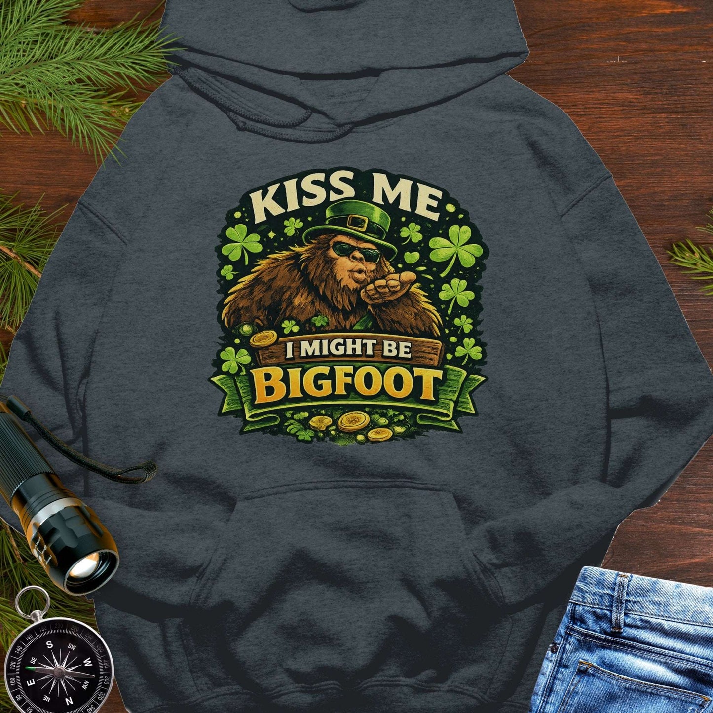 Dark Heather Gray hoodie with Bigfoot blowing a kiss, surrounded by shamrocks and gold coins, wearing a leprechaun hat, with the text “Kiss Me, I Might Be Bigfoot”