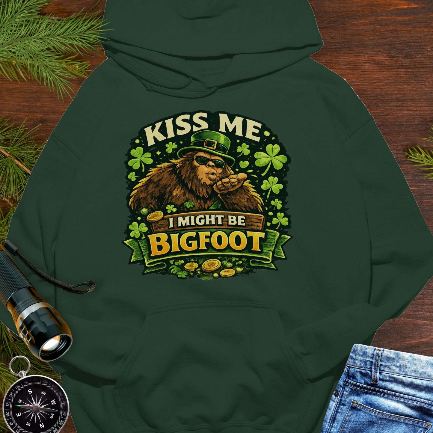 Green hoodie with Bigfoot blowing a kiss, surrounded by shamrocks and gold coins, wearing a leprechaun hat, with the text “Kiss Me, I Might Be Bigfoot”.
