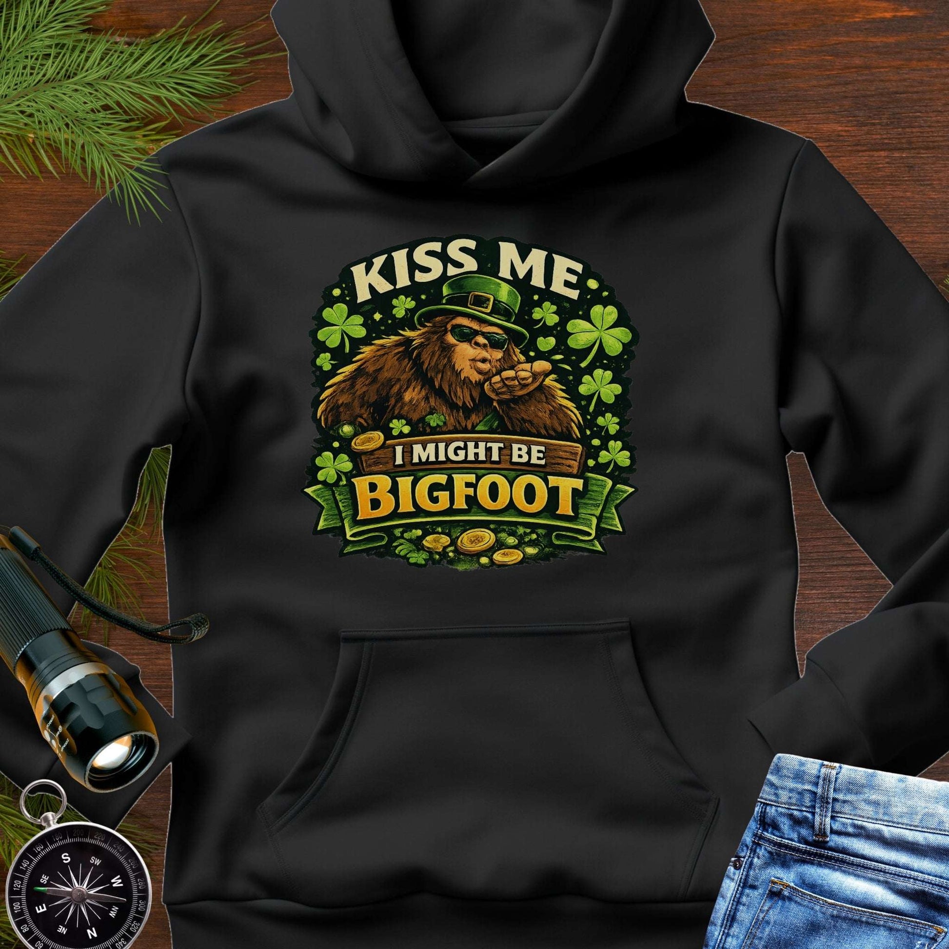 Black hoodie with Bigfoot blowing a kiss, surrounded by shamrocks and gold coins, wearing a leprechaun hat, with the text “Kiss Me, I Might Be Bigfoot”
