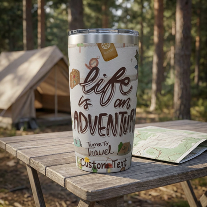 Tumbler with 'Life is an Adventure' text on a wooden table outdoors with a tent and map in the background.