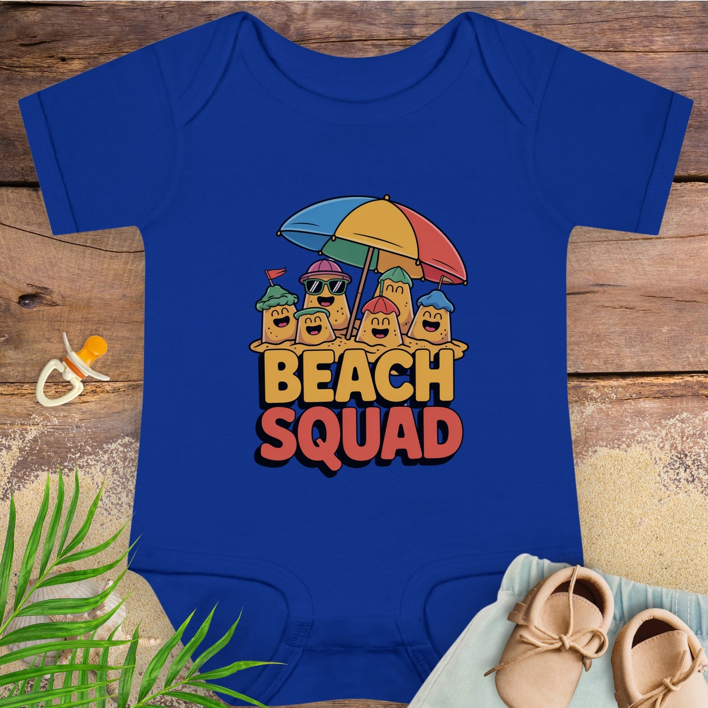 Blue baby onesie with 'Beach Squad' design on a wooden surface with beach-themed items.