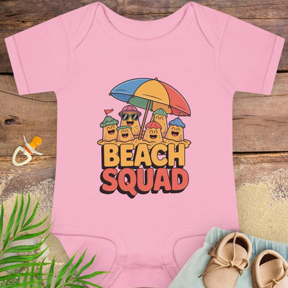 Pink baby onesie with 'Beach Squad' design on a wooden surface with beach accessories.