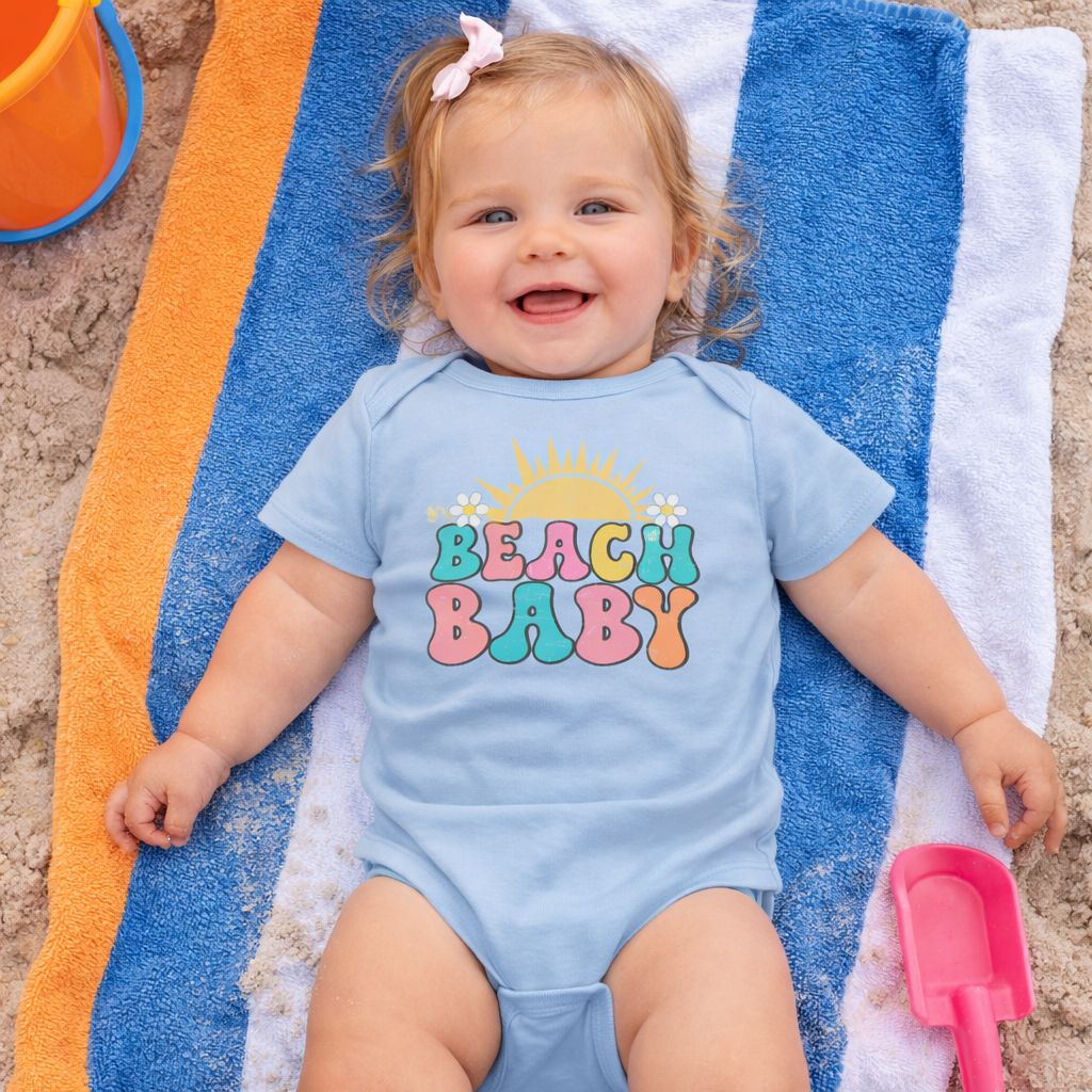 Baby wearing a 'Beach Baby' onesie on a towel with beach toys.