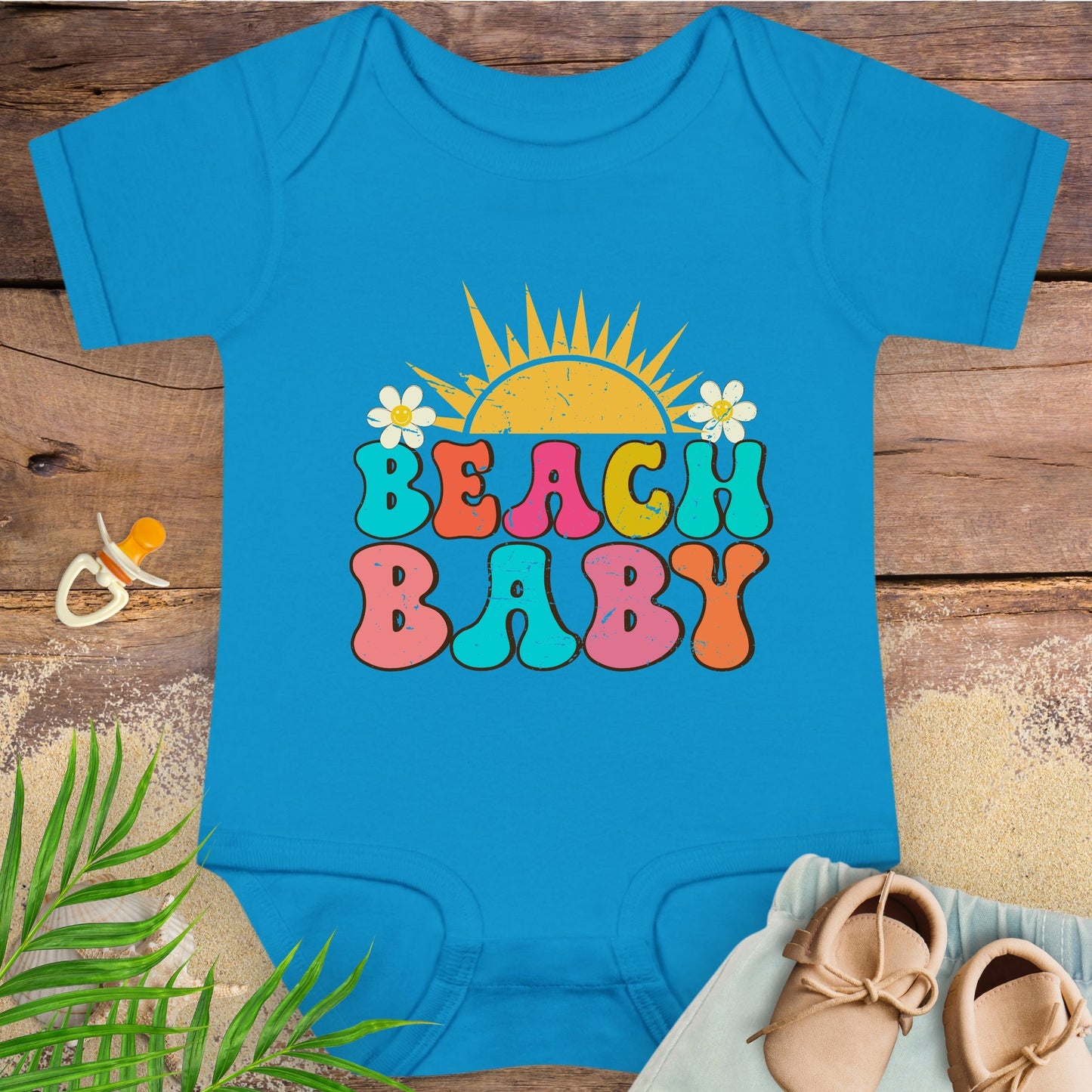 Blue baby onesie with 'Beach Baby' print on a wooden surface with beach accessories.