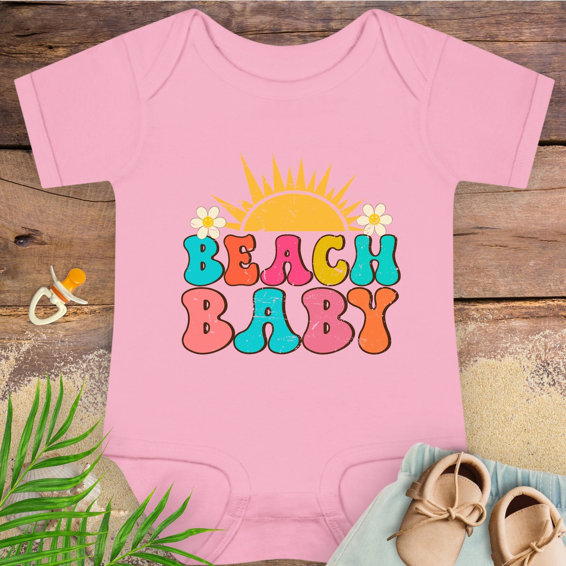 Pink baby onesie with 'Beach Baby' text on a wooden surface with beach-themed items.