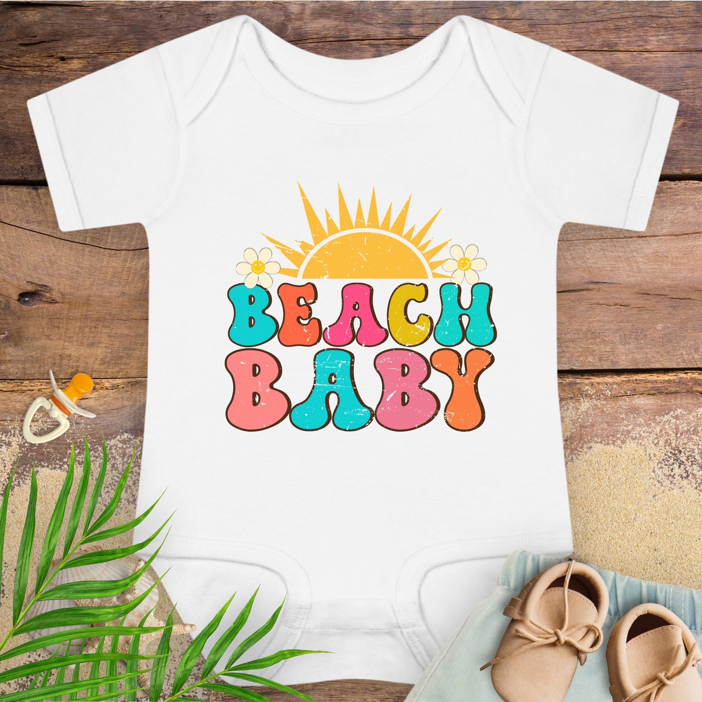 White baby onesie with 'Beach Baby' print on a wooden surface with a pacifier and sandals.