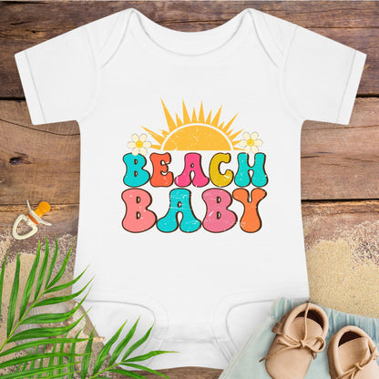 White baby onesie with 'Beach Baby' print on a wooden surface with a pacifier and sandals.