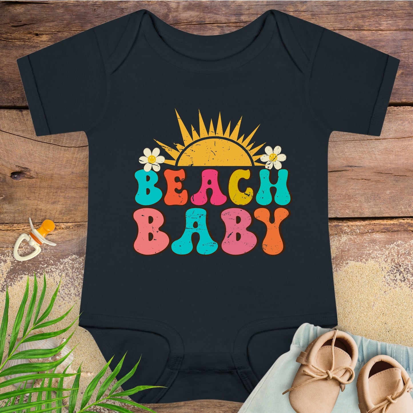 Black baby onesie with 'Beach Baby' text on a wooden surface with a pacifier and shoes.