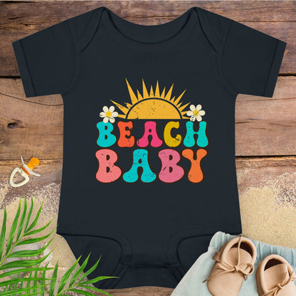Black baby onesie with 'Beach Baby' text on a wooden surface with a pacifier and shoes.