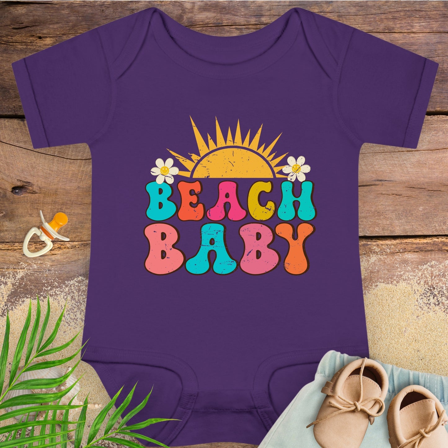 Purple baby onesie with 'Beach Baby' print on a wooden surface with a pacifier and shoes.