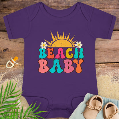 Purple baby onesie with 'Beach Baby' print on a wooden surface with a pacifier and shoes.