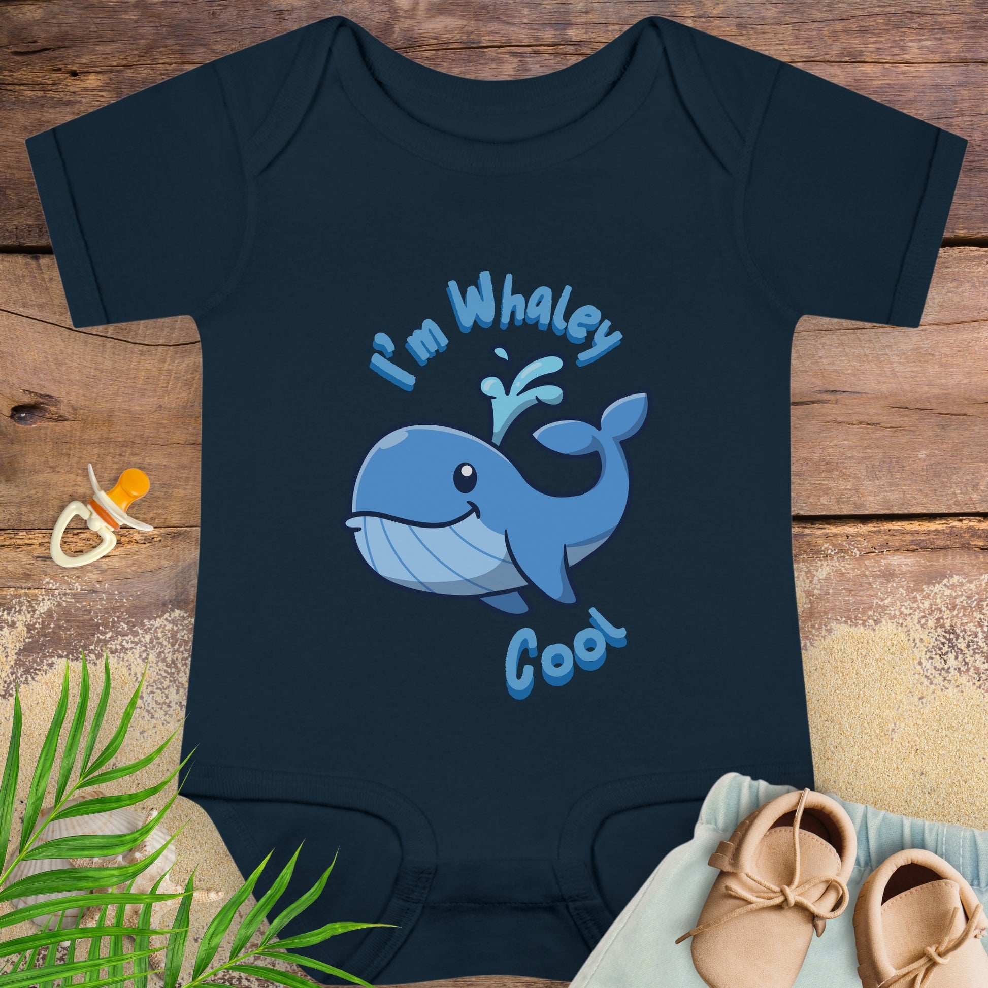 Navy blue baby onesie with a whale design and text on a wooden surface with a pacifier and shoes.