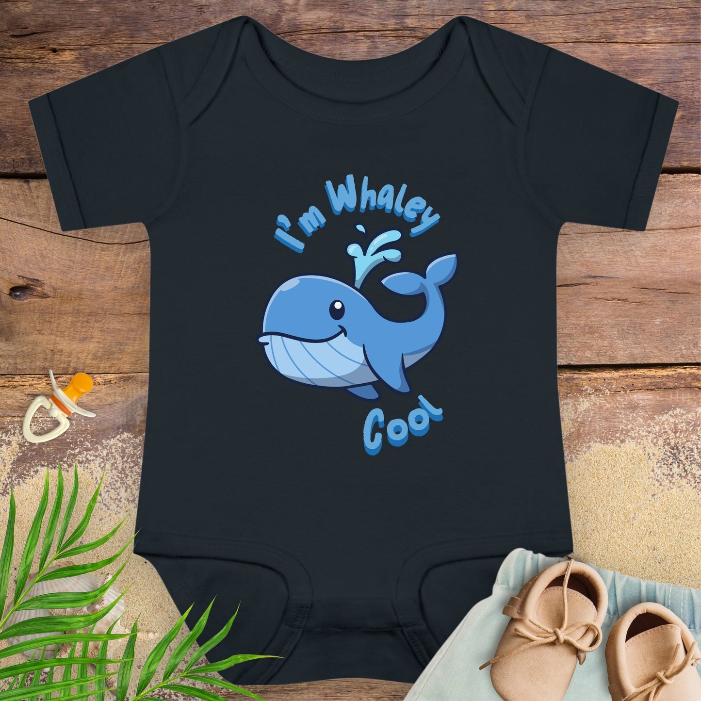 Black baby onesie with a blue whale design and text on a wooden surface with a pacifier and shoes.