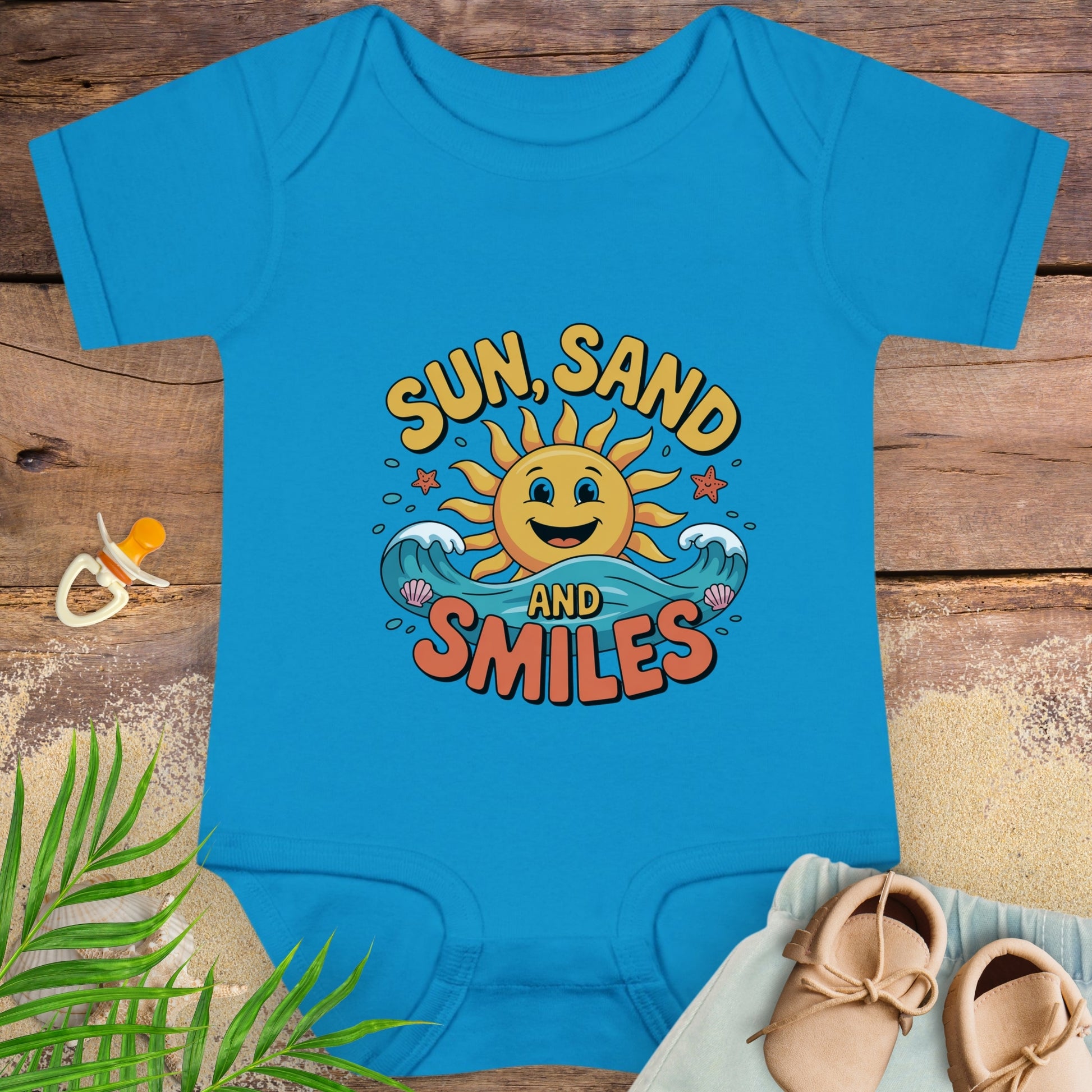 Blue baby onesie with 'Sun, Sand and Smiles' design on a wooden surface with a pacifier and shoes.
