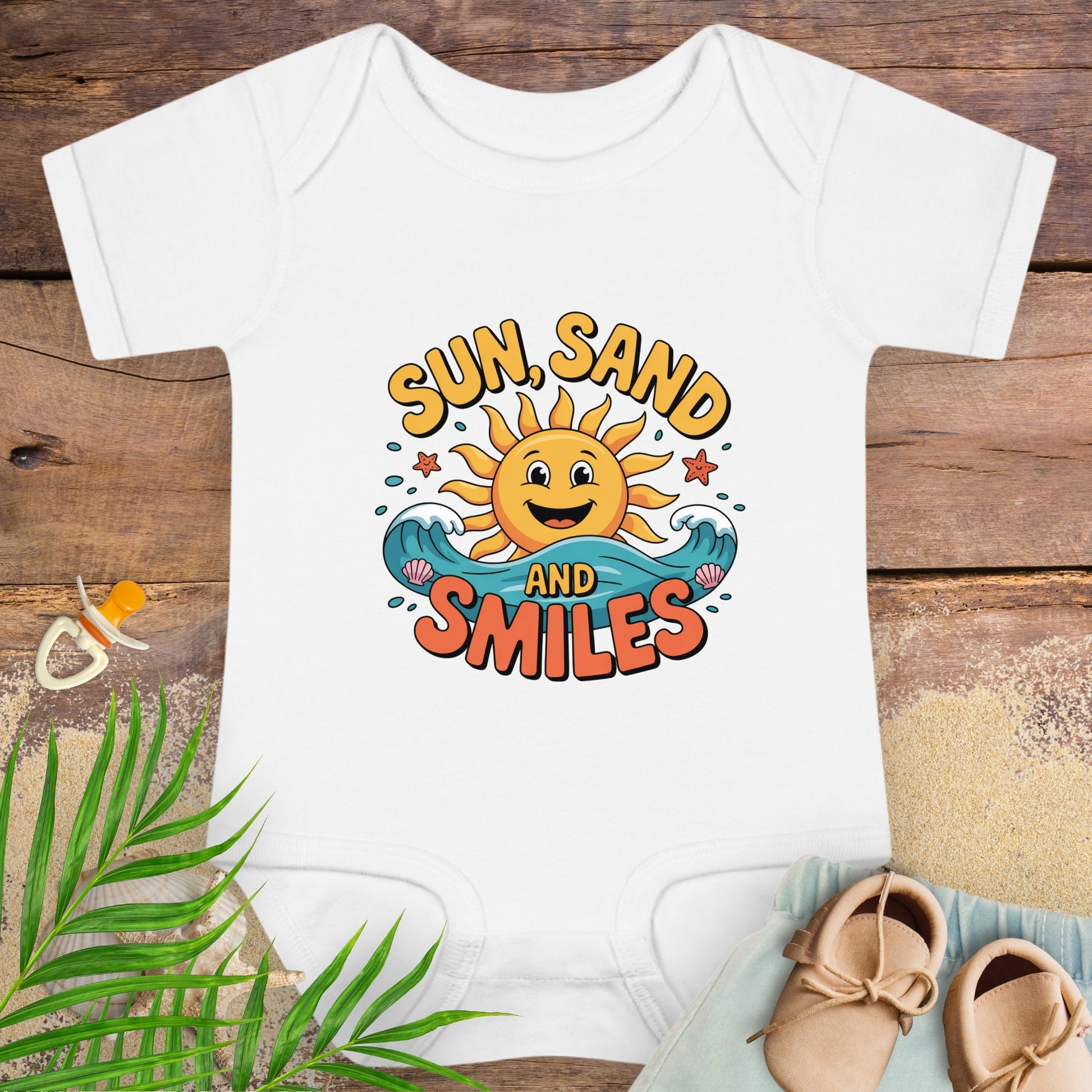 White baby onesie with 'Sun, Sand and Smiles' design on a wooden surface with beach-themed items.