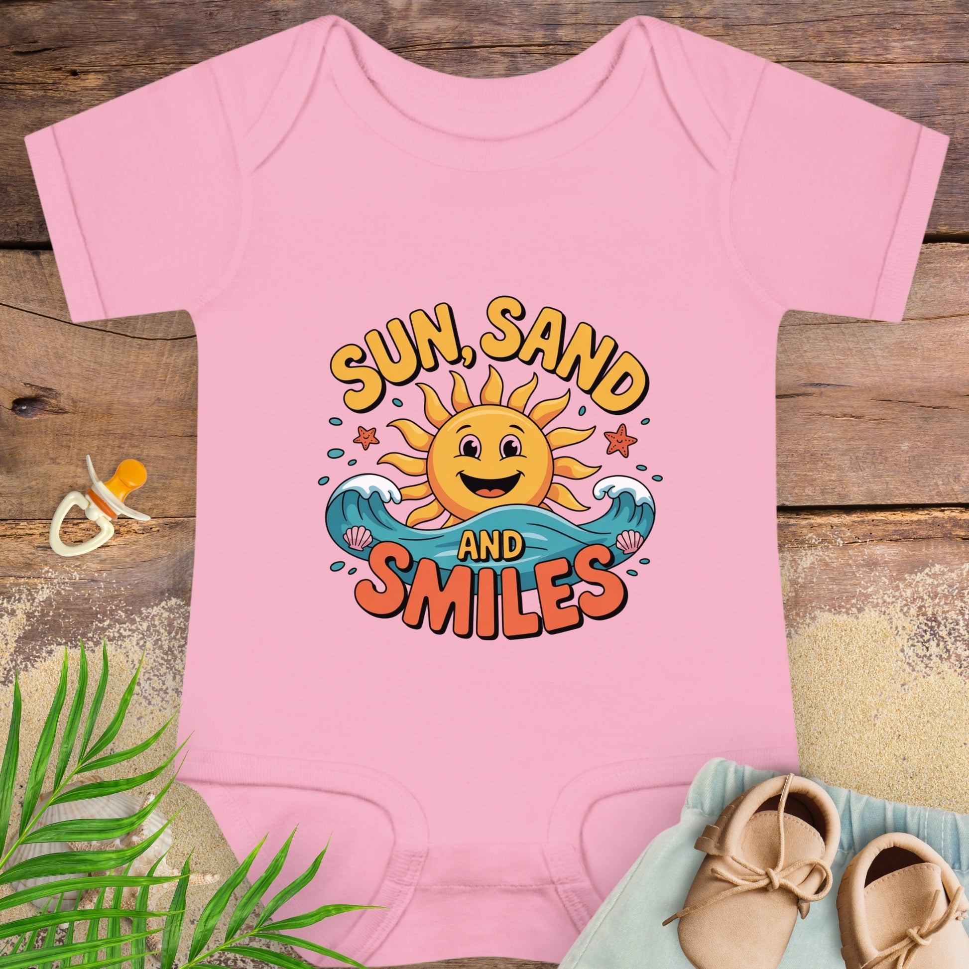 Pink baby onesie with 'Sun, Sand and Smiles' design on a wooden surface with beach-themed items.
