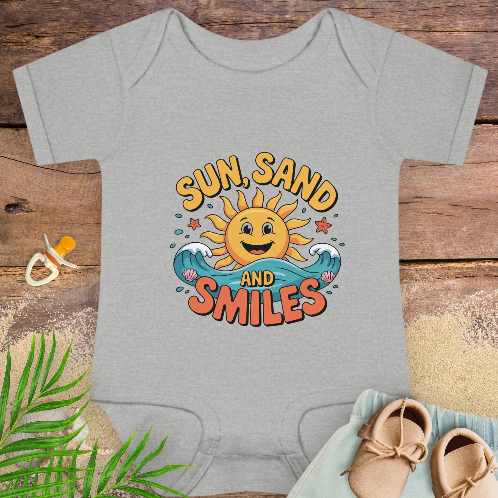 Gray baby onesie with 'Sun, Sand and Smiles' design on a wooden surface with baby items.