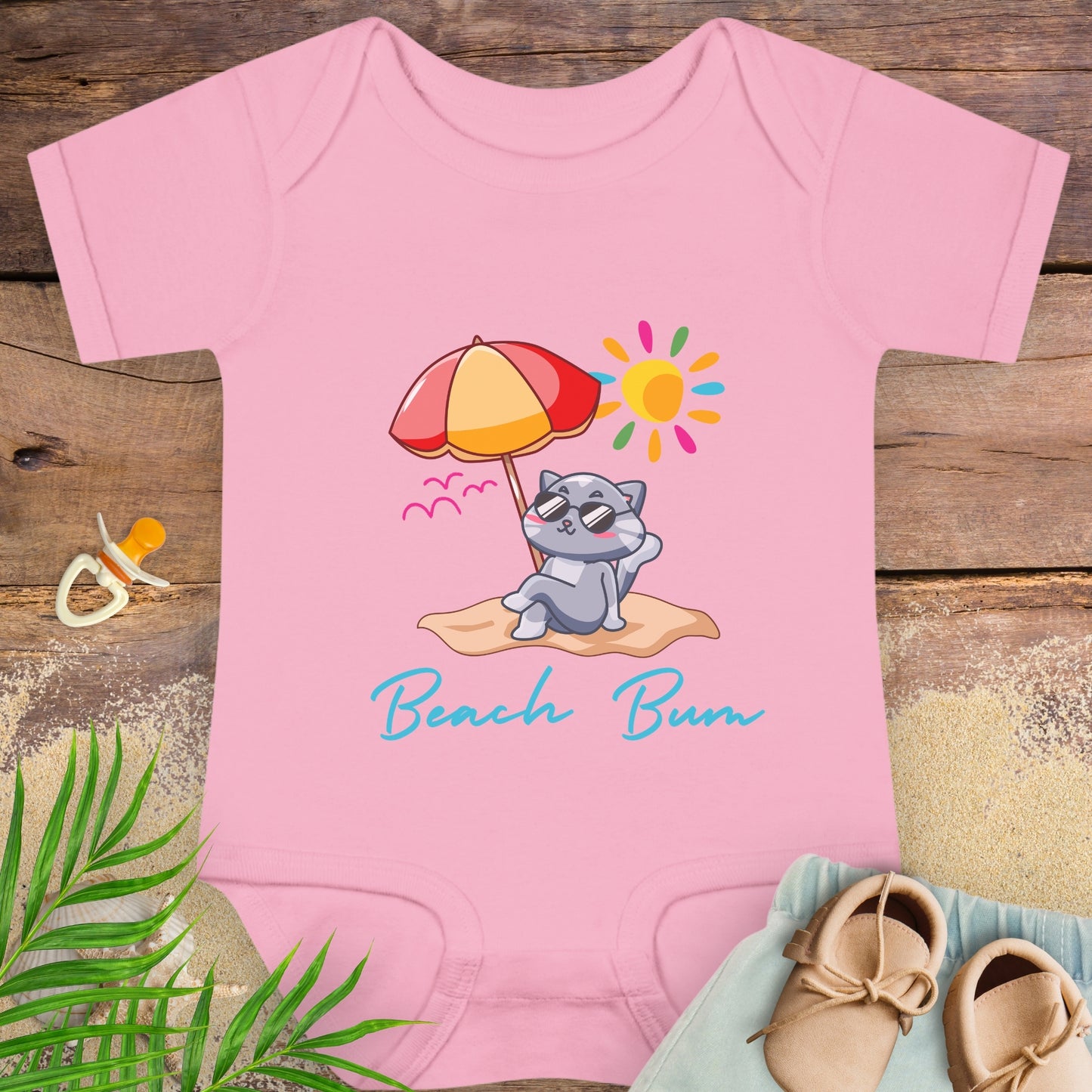 Pink baby onesie with a beach-themed design featuring a cat under an umbrella, sitting on sand under the sun.