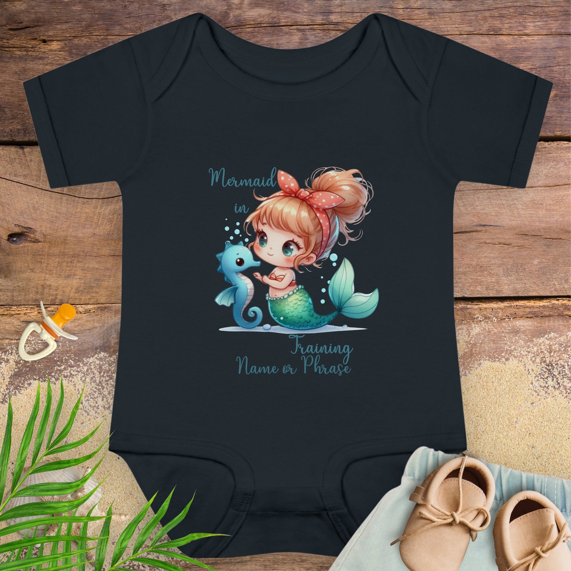 Black baby onesie with mermaid design on a wooden surface with shoes and leaves.