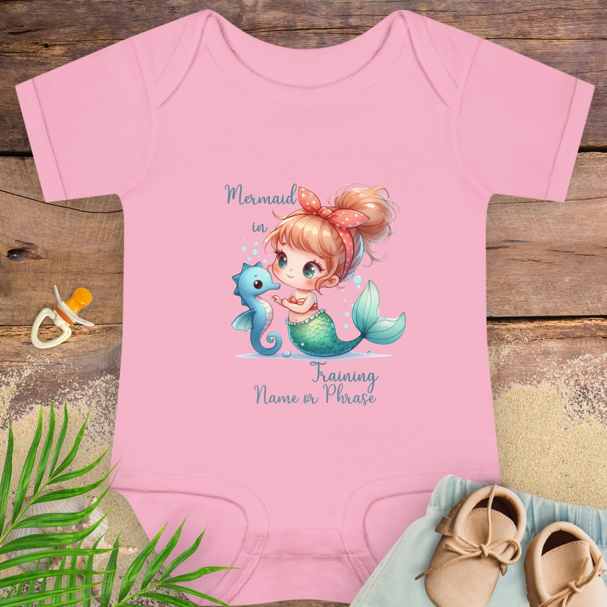 Pink baby onesie with mermaid design on a wooden surface with baby items.