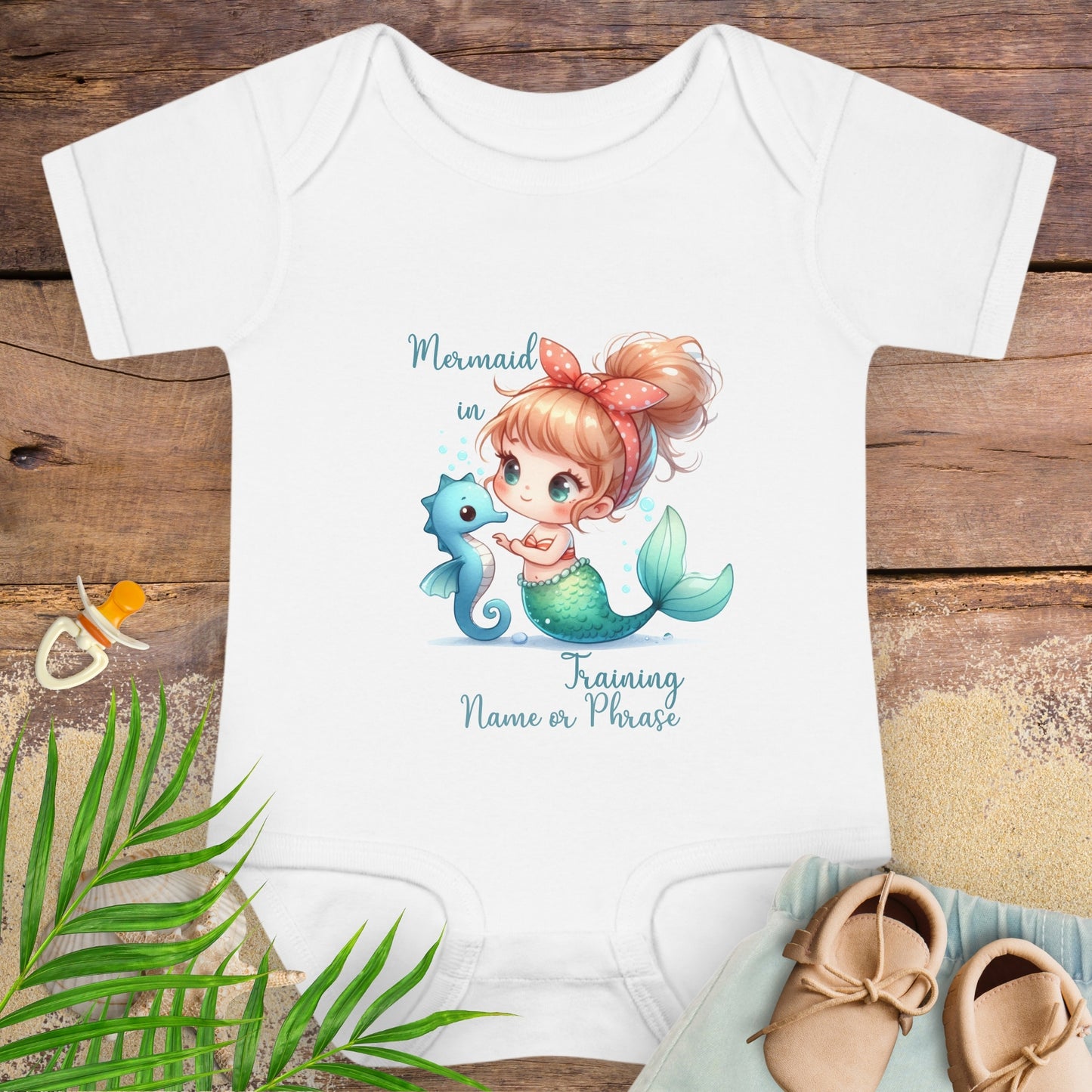 Baby onesie with mermaid design on a wooden surface with baby items.