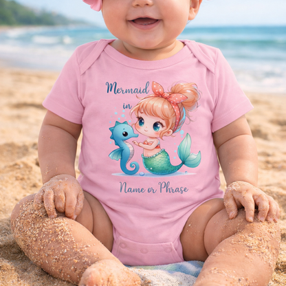 Baby wearing a pink onesie with a mermaid design on a beach.