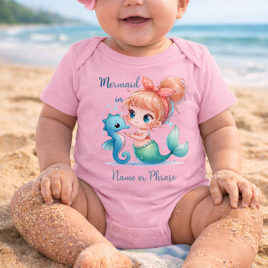 Baby wearing a pink onesie with a mermaid design on a beach.