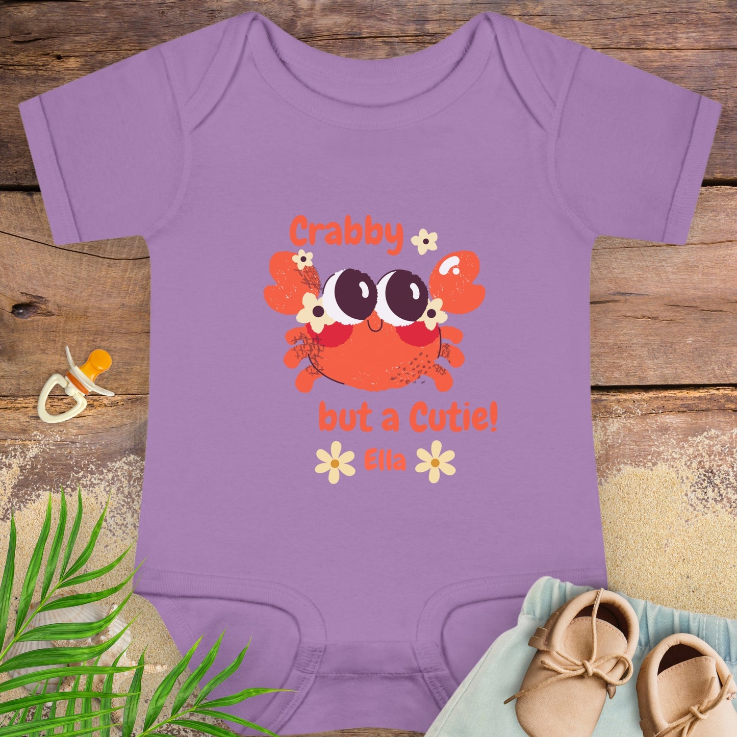 Purple baby onesie with a crab design and text on a wooden surface with shoes and a pacifier.
