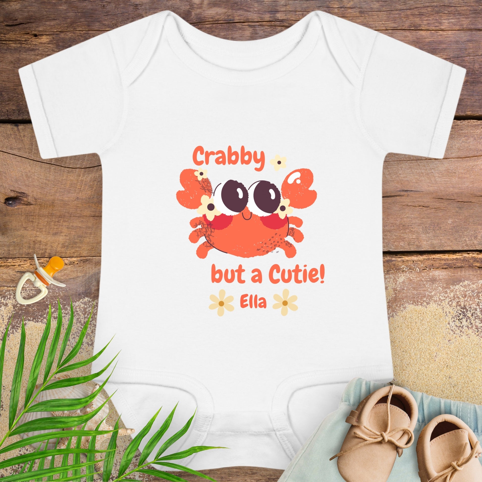 White baby onesie with a crab design and text on a wooden surface with baby items.