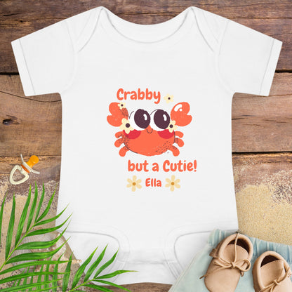 White baby onesie with a crab design and text on a wooden surface with baby items.