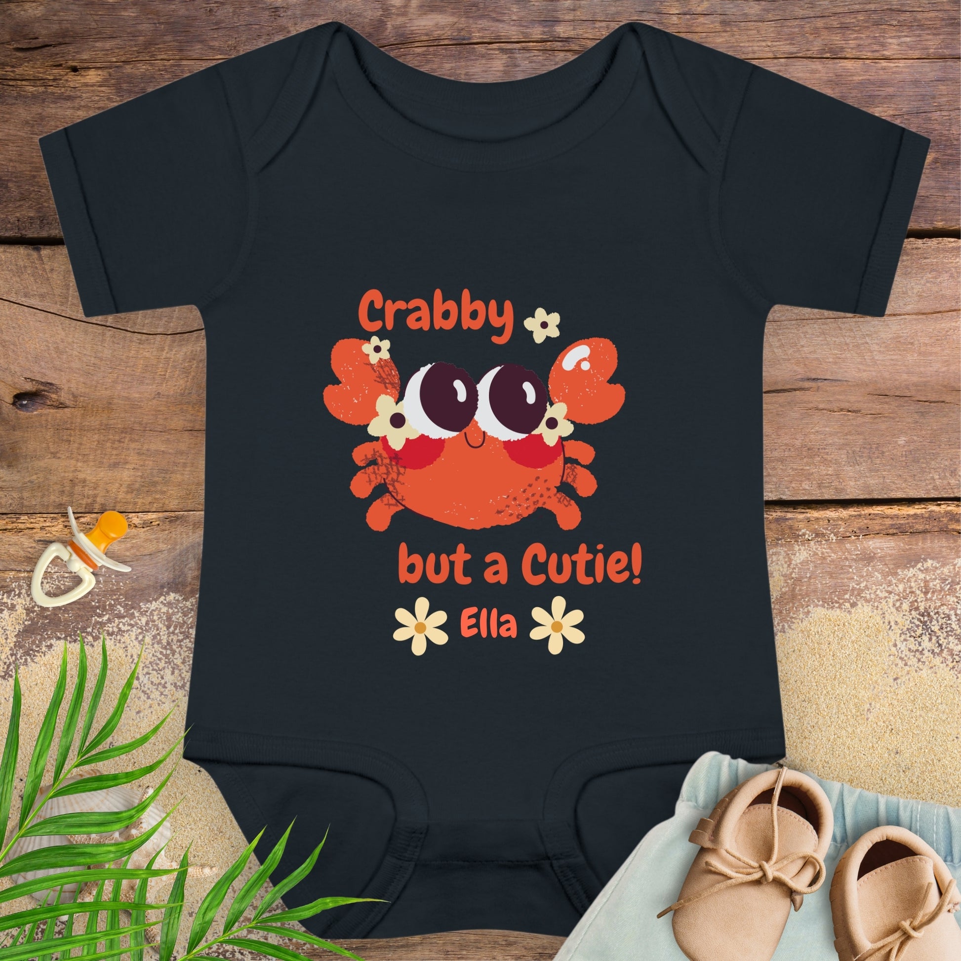 Black baby onesie with a crab design and text on a wooden surface with a pacifier and shoes.