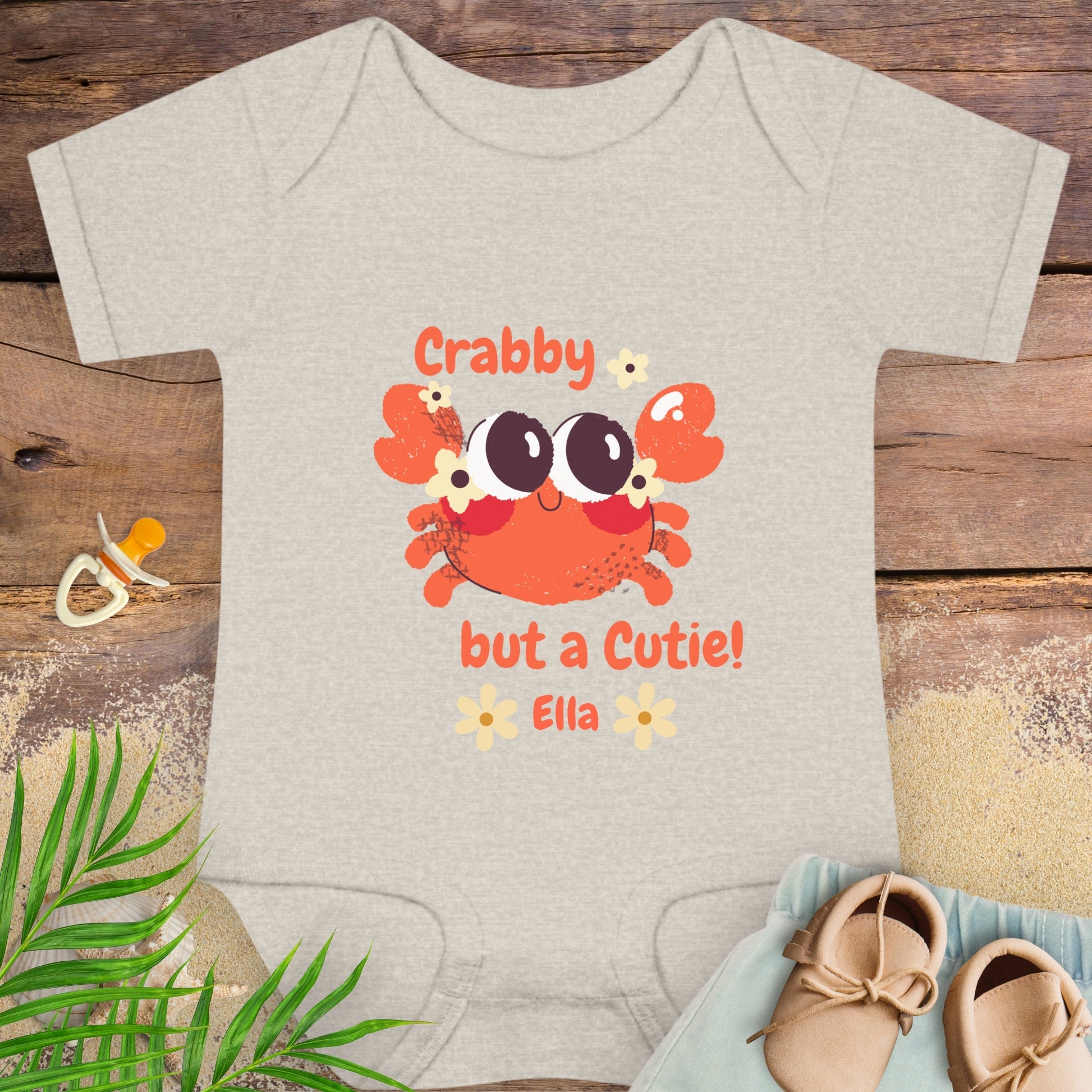 Baby onesie with a crab design and text on a wooden surface with baby items.