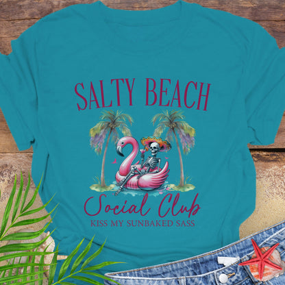 Teal t-shirt with 'Salty Beach Social Club' design featuring a flamingo and skeleton, on a wooden surface with palm leaves and starfish.